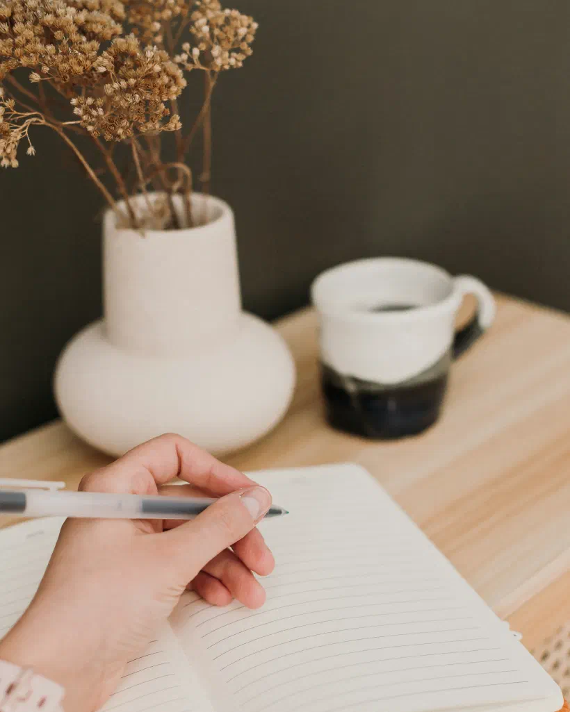 A person writing notes by hand in a notebook beside a cup of coffee